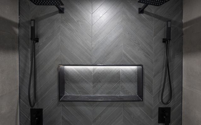 Dark chevron porcelain tile shower with dual rain heads and a lit niche