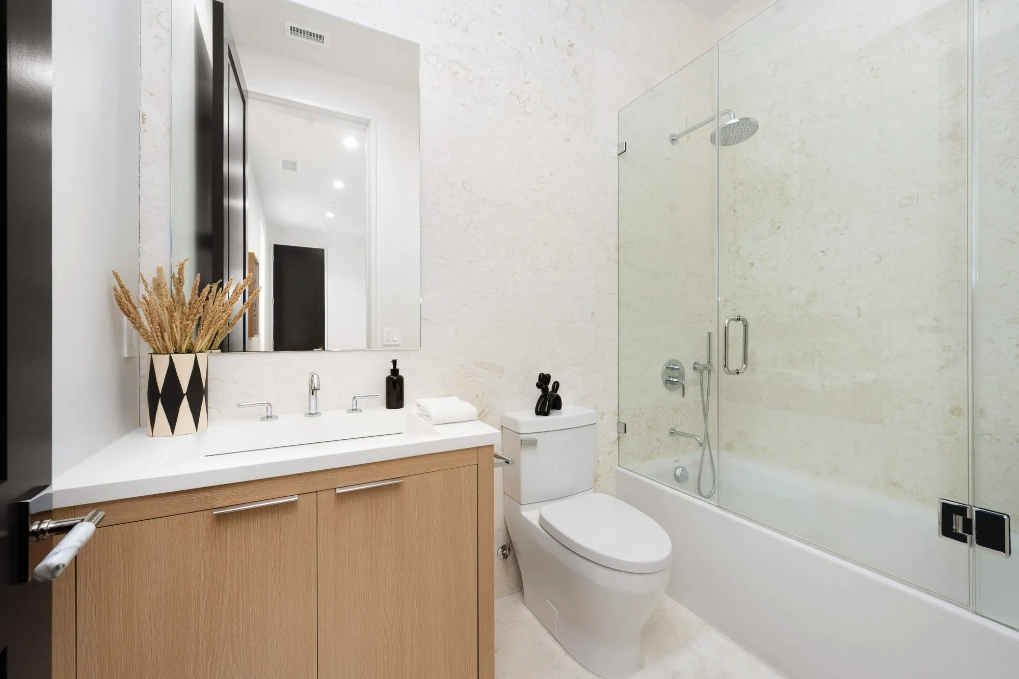 Bathroom with cream stone walls, a light oak vanity and a glass-enclosed tub shower