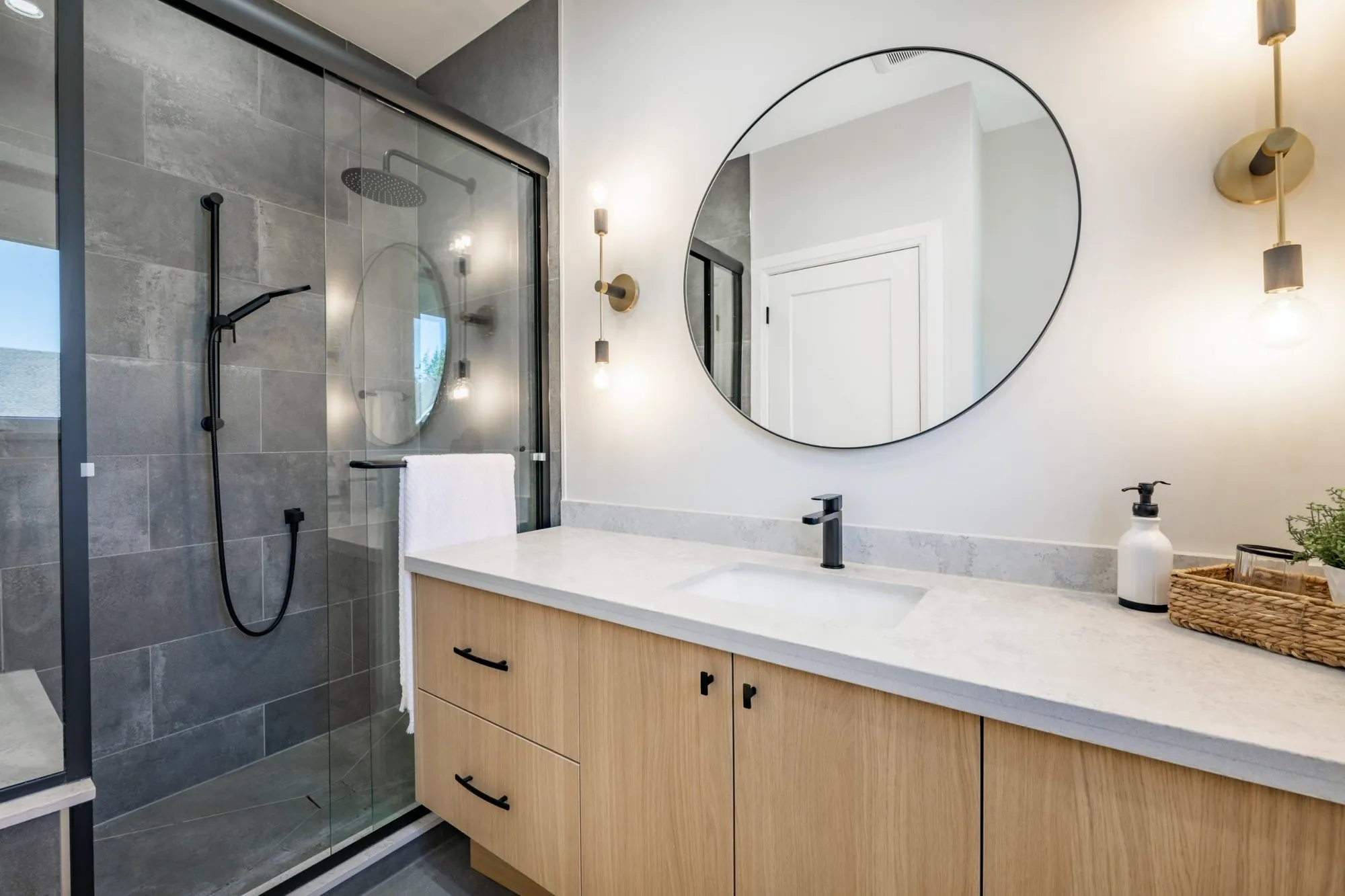 Bathroom with a light oak vanity, quartz top, round black mirror and a grey tile shower with sliding glass