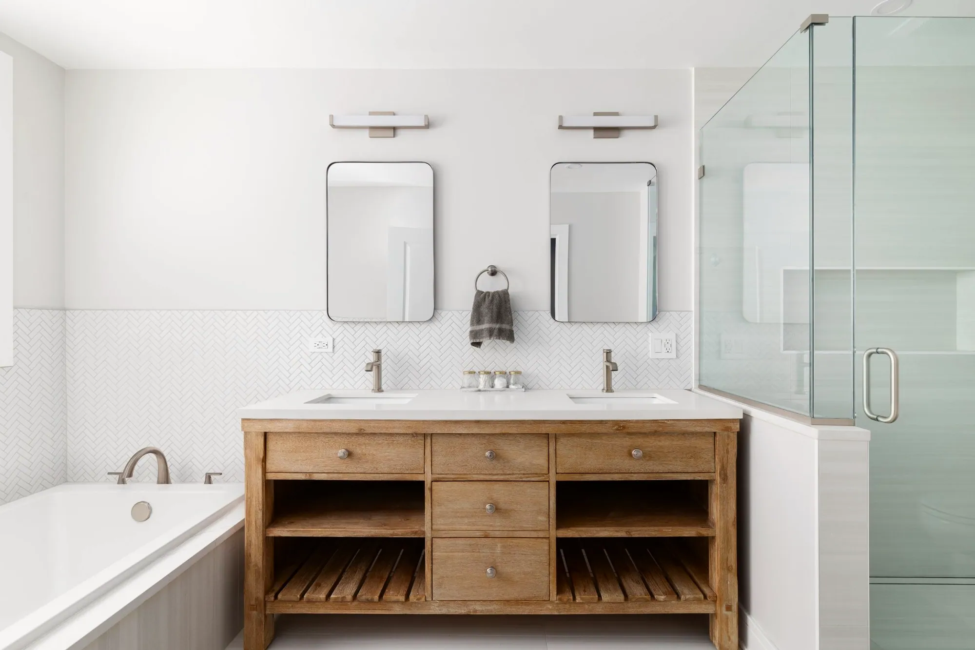 Bathroom with a light wood double vanity, white herringbone tile wainscot, soaker tub and glass shower