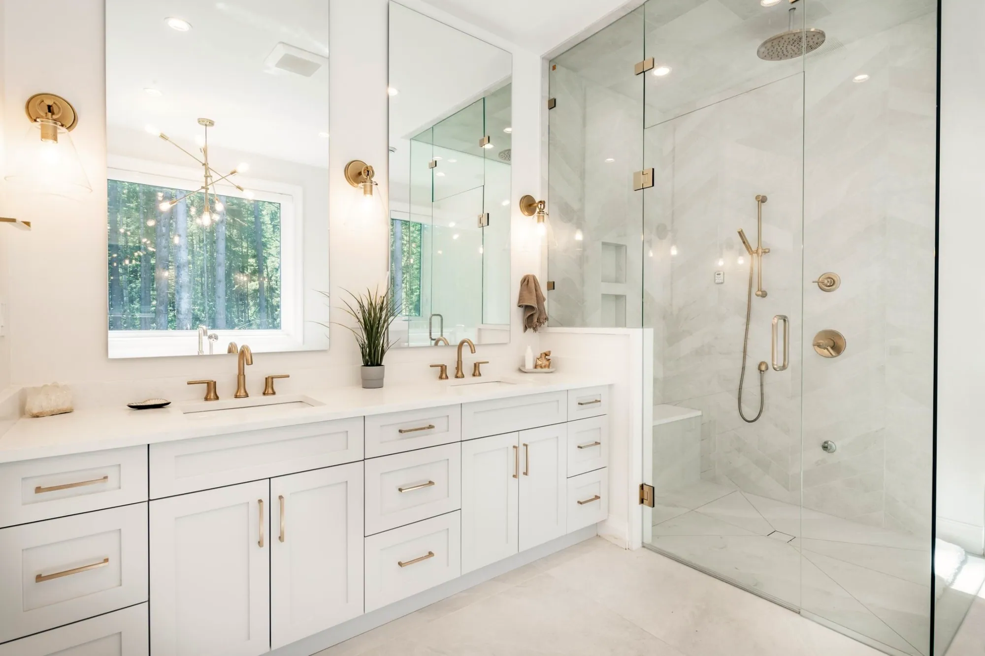 Bright ensuite with a white shaker double vanity, brushed gold faucets and sconces, and a frameless glass walk-in shower with a bench