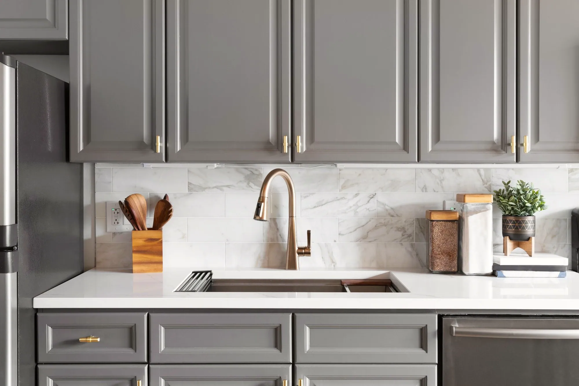 Grey painted kitchen cabinets with brass hardware, a marble-look subway backsplash and a bronze faucet