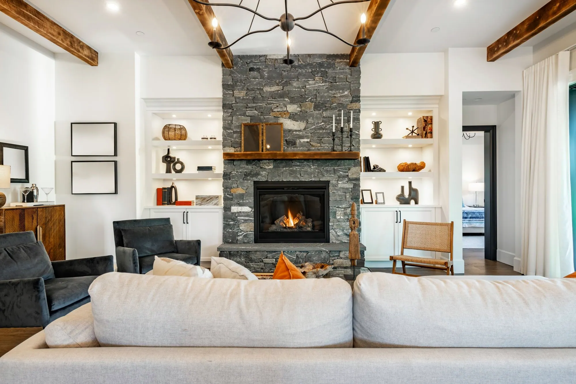 Living room with a stacked stone fireplace, wood mantel, lit built-in shelves and wood ceiling beams