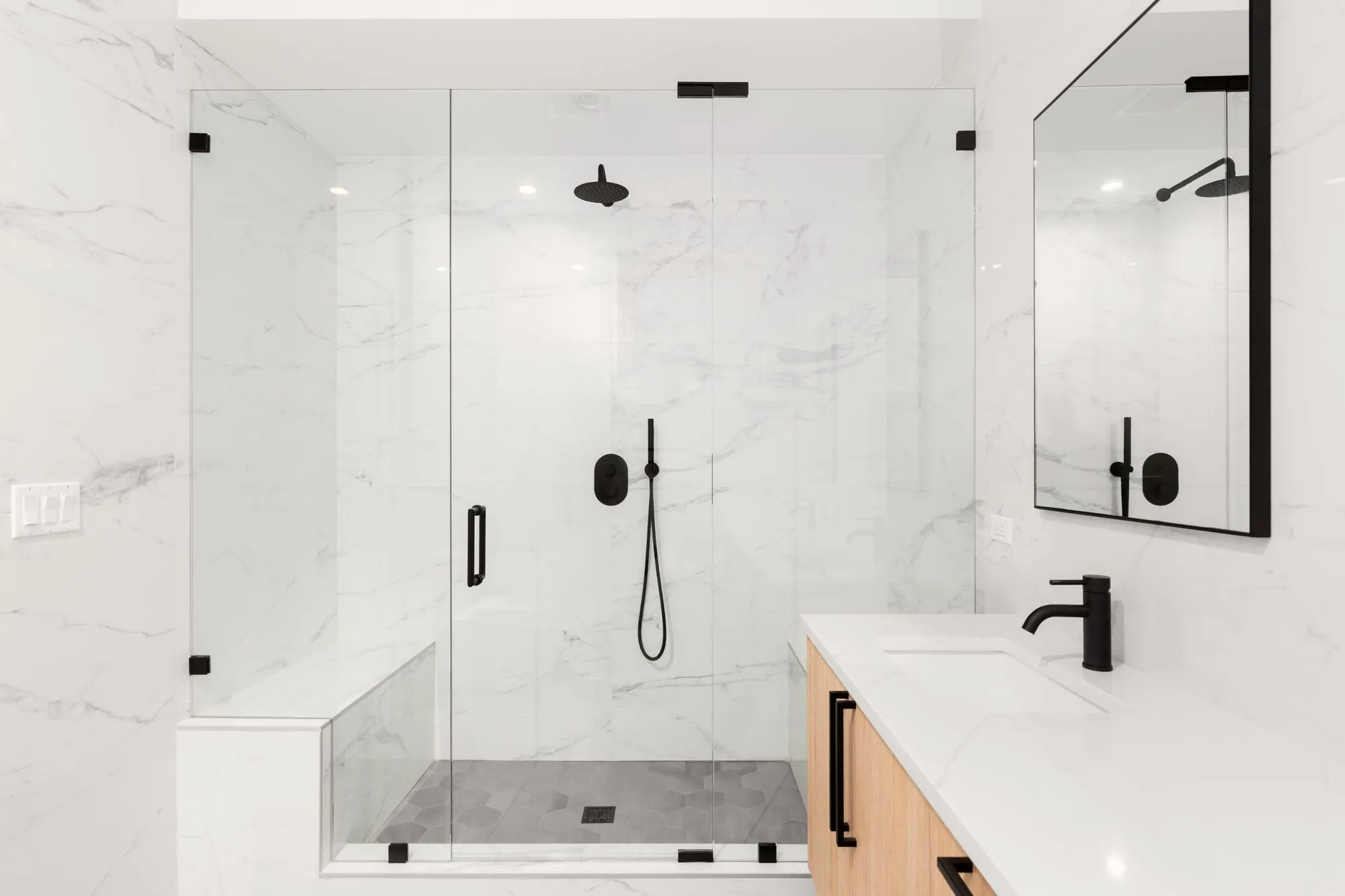 Marble-look tile bathroom with a large glass shower and bench, black fixtures and a light wood vanity