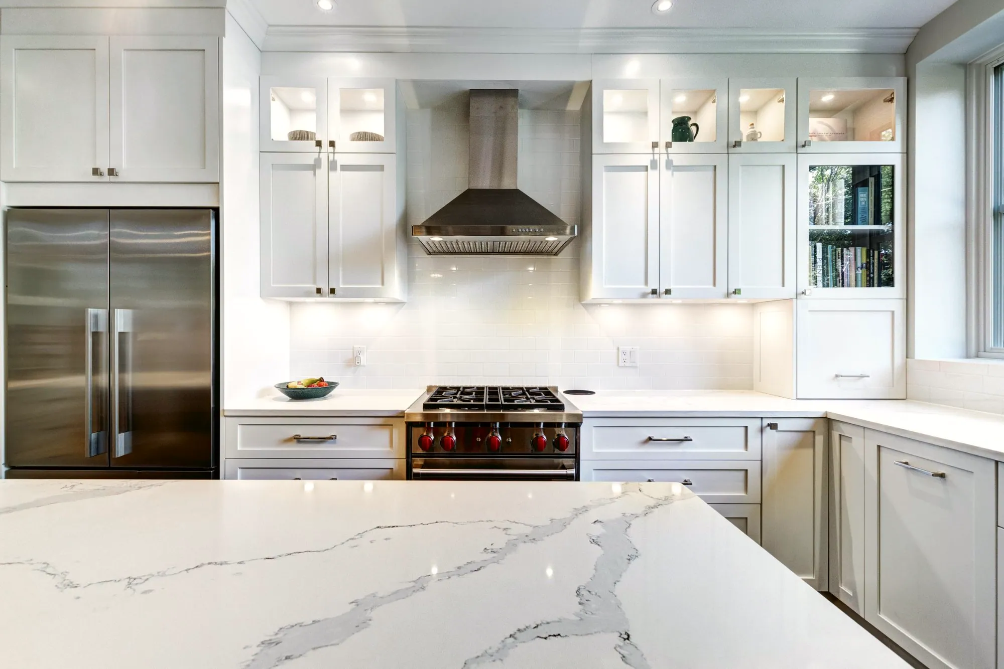 White kitchen with glass-front upper cabinets, a veined quartz island and a range with red knobs